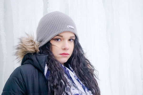 A person wearing a gray beanie and a black coat with a fur-trimmed hood stands in front of an icy background.