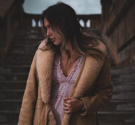 A person with long hair wearing a tan shearling coat and a light-colored top stands on a staircase, looking down to the side.