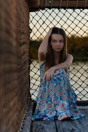 A person with long hair sits on a wooden surface, wearing a patterned blue dress, with a hand on their head. A wire fence and water are in the background.