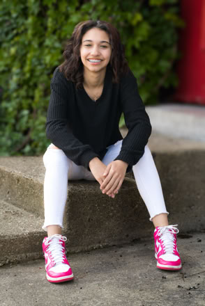 A person with curly hair sits on a concrete step, wearing a black long-sleeve shirt, white pants, and pink sneakers, with greenery and a red wall in the background.