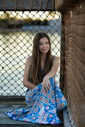 A person with long hair sitting on a wooden surface in front of a chain-link fence, wearing a blue patterned dress.