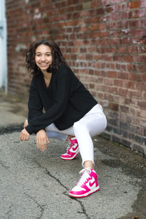 A person squatting on a sidewalk, wearing a black sweater, white pants, and pink sneakers, with a brick wall in the background.