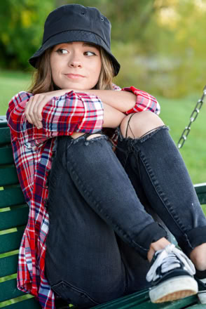A person wearing a black bucket hat, red plaid shirt, and ripped jeans sits on a green swing, with knees drawn up and arms wrapped around them.