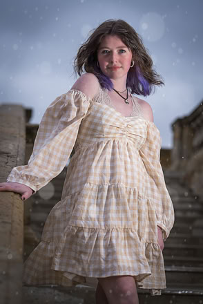 A person with long hair and purple highlights wearing a checkered off-shoulder dress stands on stone steps, with a cloudy sky in the background.
