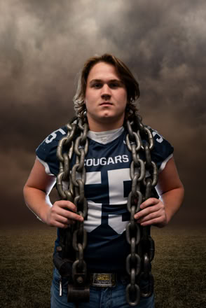 A person wearing a football jersey with the number 55 and the word "Cougars" holds large chains around their shoulders, standing against a cloudy sky background.