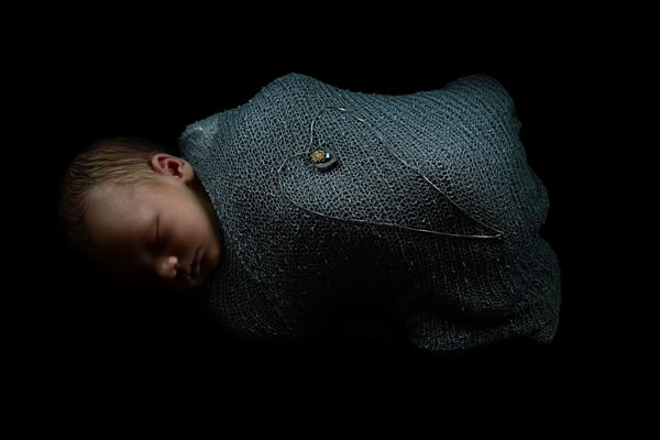 A baby wrapped in a textured blue blanket, sleeping on a dark background.