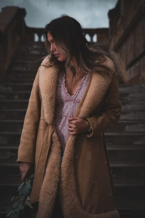 A person wearing a long, tan shearling coat and a light-colored dress stands on outdoor steps, holding a small bouquet of flowers, with a stone railing in the background.