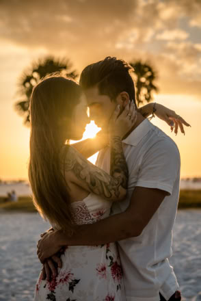 A couple embracing on a beach at sunset, with palm trees in the background.